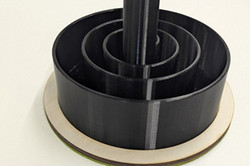 Ein runder, schwarzer Organizer aus dem 3D-Drucker mit mehreren konzentrischen Fächern und einem hohen Mittelrohr steht auf einer Holzplatte. Seitlich ist ein kleiner Griff angebracht, an dem kleinere Gegenstände aufgehängt werden können..