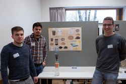 Drei Studierende der RWTH Aachen stehen an einem Tisch, auf dem sie ein Modell für einen Wasserfilter aus zwei Getränkeflaschen aufgebaut haben.