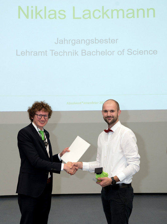 Zwei Männer schütteln sich bei einer feierlichen Ehrung die Hände. Der Mann links, Dekan Prof. Schulze-Darup, mit lockigen Haaren und Brille, trägt einen Anzug und überreicht ein weißes Zertifikat. Der Mann rechts, Niklas Lackmann, mit Bart und roter Fliege, nimmt das Zertifikat entgegen und hält in der anderen Hand eine kleine Trophäe. Im Hintergrund wird auf einer Leinwand der Text "Niklas Lackmann, Jahrgangsbester, Lehramt Technik Bachelor of Science" angezeigt.