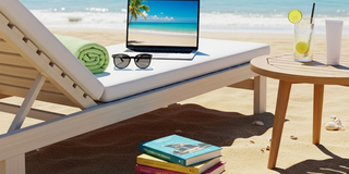 Eine Holzliege steht an einem sonnigen Sandstrand, im Hintergrund das glitzernde blaue Meer. Auf der Liegefläche liegen ein zusammengerolltes, hellgrünes Handtuch, eine schwarze Sonnenbrille und ein offener Laptop, dessen Bildschirm einen Strand zeigt. Vor der Liege sind Bücher im Sand gestapelt. Rechts neben der Liege steht ein kleiner Holztisch, auf dem ein Glas mit einem kühlen Getränk und einer Limettenscheibe steht. Die Szene ist von hellem Sonnenlicht erfüllt.