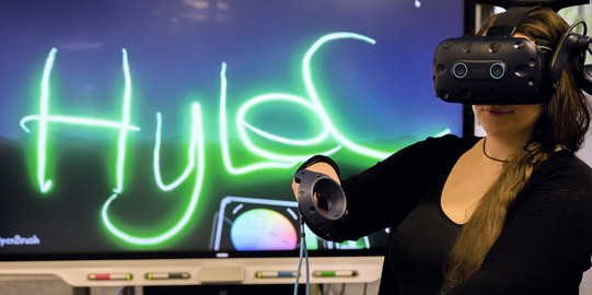 Das Bild zeigt eine junge Frau mit einer VR-Brille vor einem großen Monitor