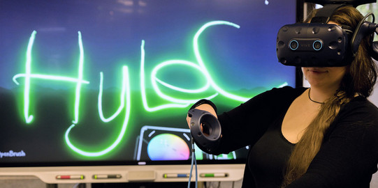 Das Bild zeigt eine junge Frau mit einer VR-Brille vor einem großen Monitor