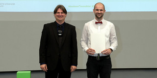 Zwei Männer posieren für ein Foto bei einer Absolventenfeier. Der Mann links, Studiendekan Prof. Tobias Haertel, trägt einen dunklen Anzug über einem dunklen Hemd. Der Mann rechts, Niklas Lackmann, trägt ein weißes Hemd, eine rote Fliege und hält ein weißes Zertifikat in den Händen. Beide lächeln in die Kamera. Im Hintergrund ist eine Leinwand mit der Aufschrift "Absolvent*innenfeier" zu sehen.