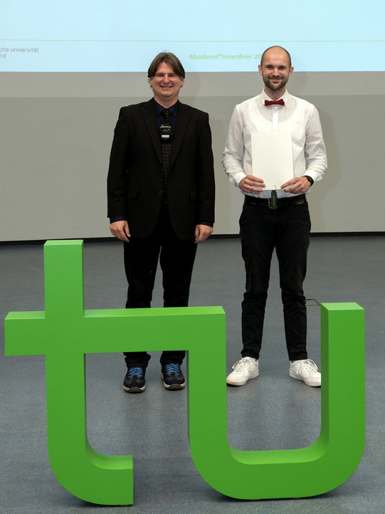 Zwei Männer posieren für ein Foto bei einer Absolventenfeier. Sie stehen hinter einem großen, leuchtend grünen Logo der Technischen Universität (tu). Der Mann links, Studiendekan Prof. Tobias Haertel, trägt einen dunklen Anzug, der Mann rechts, Niklas Lackmann, ein weißes Hemd mit roter Fliege und hält ein weißes Zertifikat in den Händen. Beide lächeln in die Kamera.
