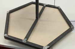 Ein zweistöckiger, sechseckiger Ständer aus Holz und geschweißten Metallprofilen. Die beiden Ablageflächen sind über eine mittige Stütze drehbar miteinander verbunden.