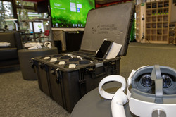 Blick über den VR-Bereich. Im vorderen Bereich liegt eine VR-Brille mit zwei Controllern, in der Mitte steht ein aufgeklappter Koffer mit weiteren VR-Brillen und im Hintergrund ist ein großer Monitor zu sehen.