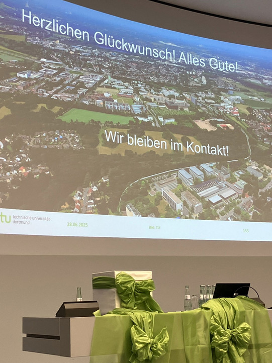 Eine Präsentationsfolie wird auf eine große Leinwand projiziert. Die Folie zeigt eine Luftaufnahme des Campus der Technischen Universität Dortmund. Darüber steht in großer, weißer Schrift: "Herzlichen Glückwunsch! Alles Gute!" und "Wir bleiben in Kontakt!". Am unteren Rand der Folie sind das Logo der TU Dortmund und das Datum 28.06.2025 zu sehen. Im Vordergrund steht ein Tisch, der mit hellgrünem, glänzendem Stoff und großen Schleifen dekoriert ist.