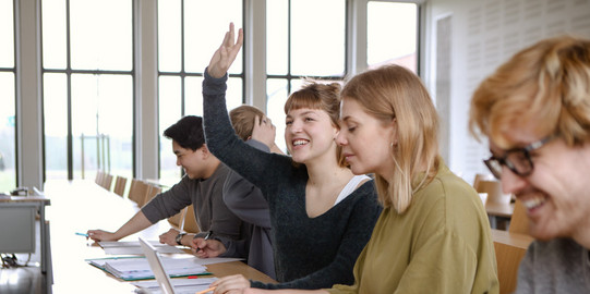 Fünf Studierende sitzen in einem Seminarraum an Tischen. Eine Studentin hebt ihre Hand.